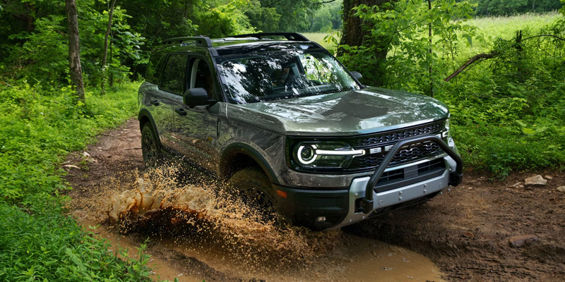 A green Ford Bronco Sport driving through muddy terrain in a forest.