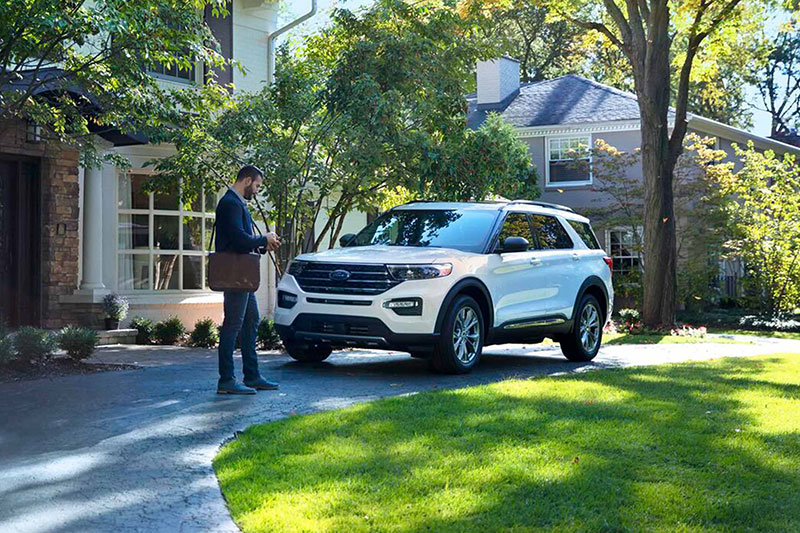 A man with a bag approaches a white Ford parked in a sunny driveway. The scene is tranquil, with green grass and trees surrounding a suburban house.