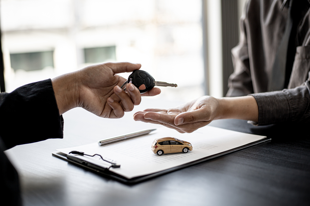 A person handing car keys to another over a desk with a miniature car and paperwork.